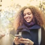 Mulher na calçada sorrindo e mexendo no celular