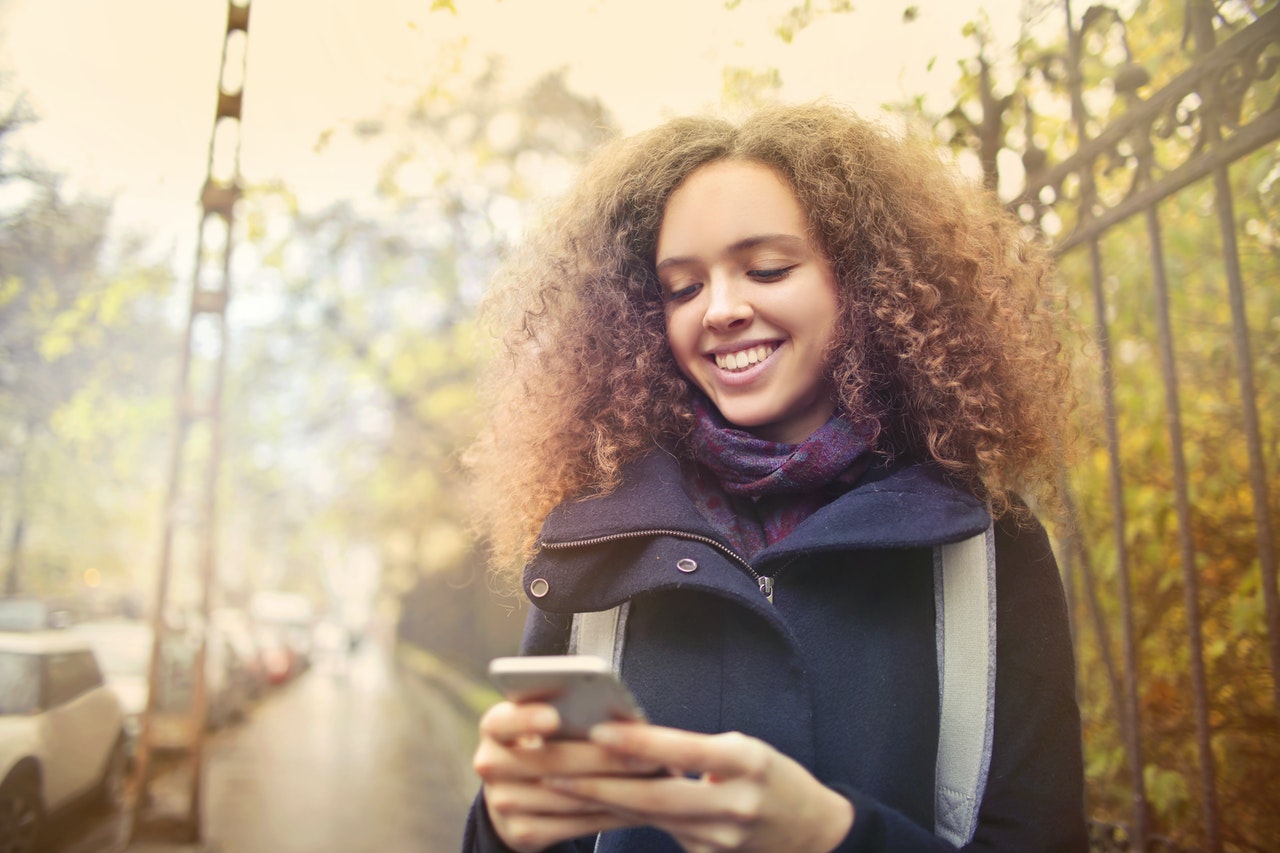 Mulher na calçada sorrindo e mexendo no celular